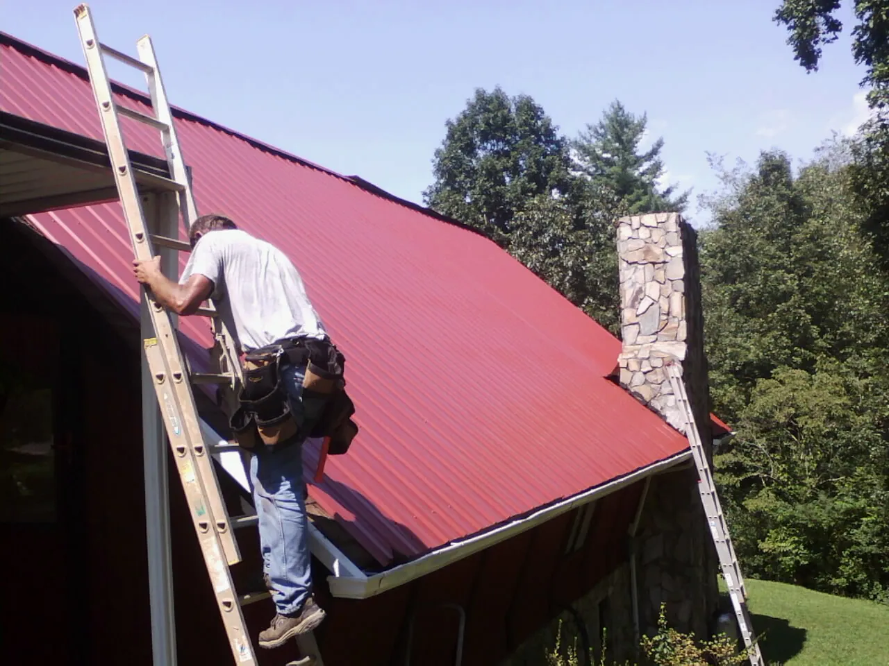 Roofer on a ladder beside a red metal roof and stone chimney — Soffit & Fascia Repair by Roofing Brothers in Voorhees